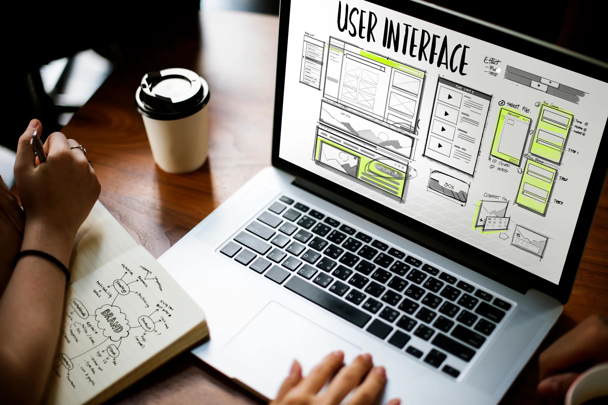Hands sketching beside a laptop displaying interface wireframes and a takeaway coffee